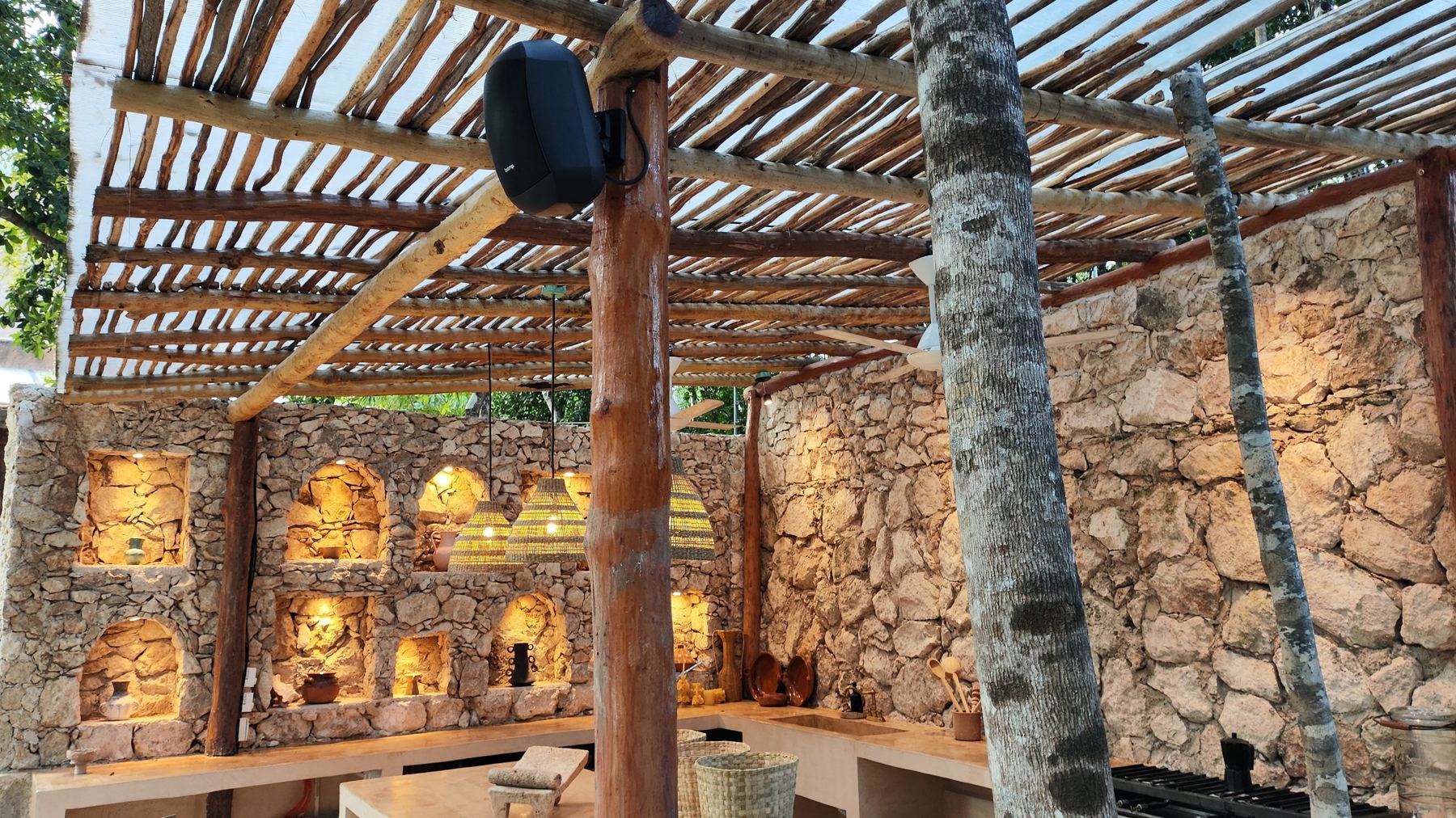 Outdoor loudspeaker under a timber pergola beside lit stone walls