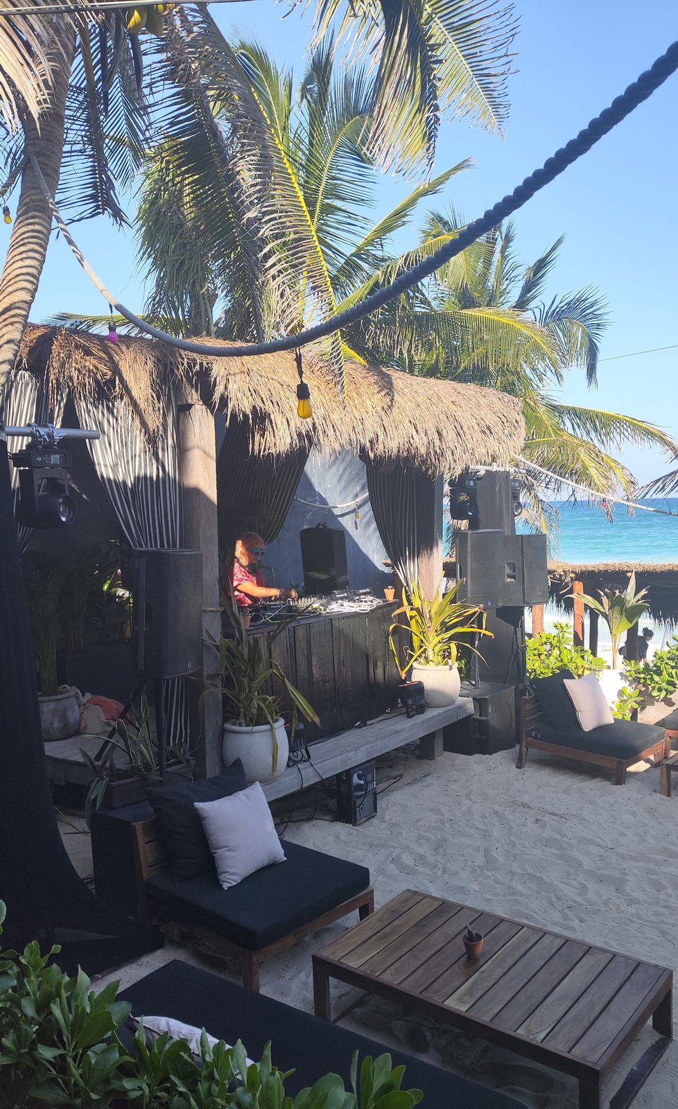 DJ in the booth with loudspeakers on each side, among palms by the sea